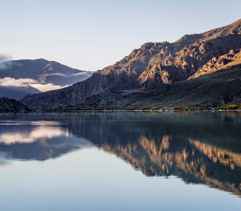 A symmetrical reflection of jagged, sunlit mountains and soft morning mist on the glassy surface of Lake Dunstan in Central Otago.