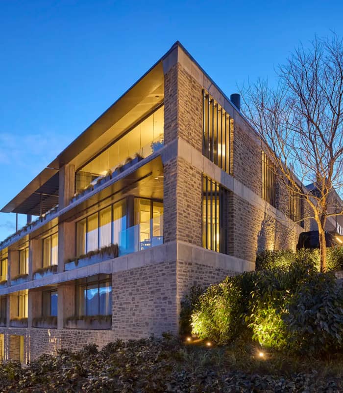 Modern stone building with large windows and lights on at dusk next to trees and bushes.