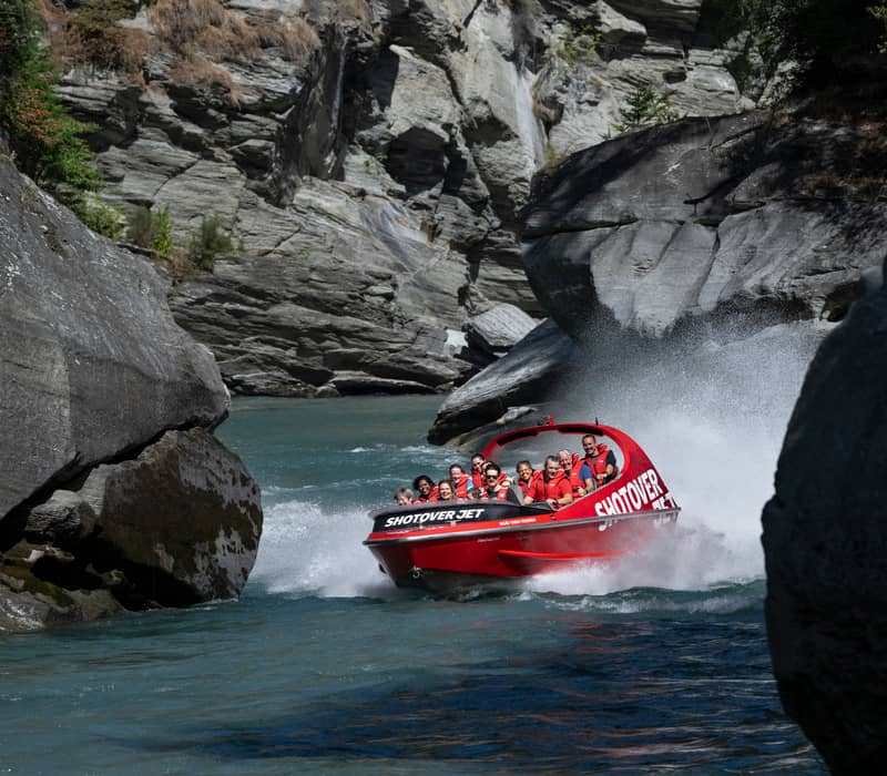 Red jet boat with tourists speeding through rocky canyon with water spray around.
