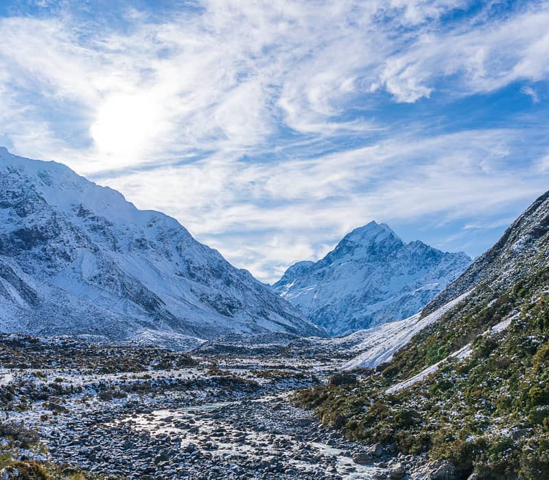 A wide view of the rocky Hooker River valley floor leading toward the massive, snow-covered peak of Aoraki / Mount Cook under a bright, wispy blue sky.