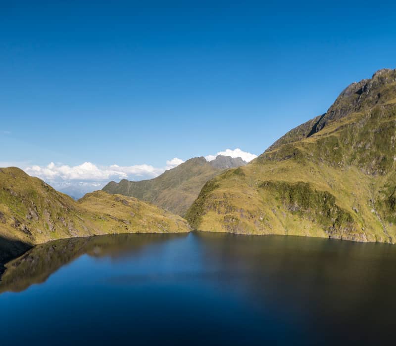 A wide panoramic view of a deep blue alpine lake surrounded by steep, sunlit golden-green mountain ridges under a clear blue sky.