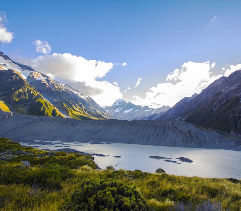A wide-angle daytime view of the Mueller Glacier lake and moraine wall, with sunlit green and gold tussock in the foreground and snow-capped peaks under a bright, cloudy sky.