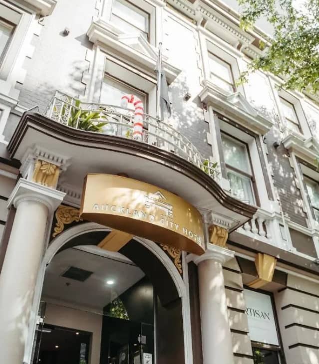 Hotel entrance with white columns, gold details, and a balcony with plants under daylight.