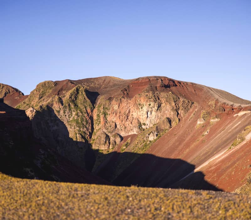 A wide view of the colorful volcanic slopes of Mount Tarawera under a clear sky, showing deep red scoria and rocky ridges in the afternoon sun.