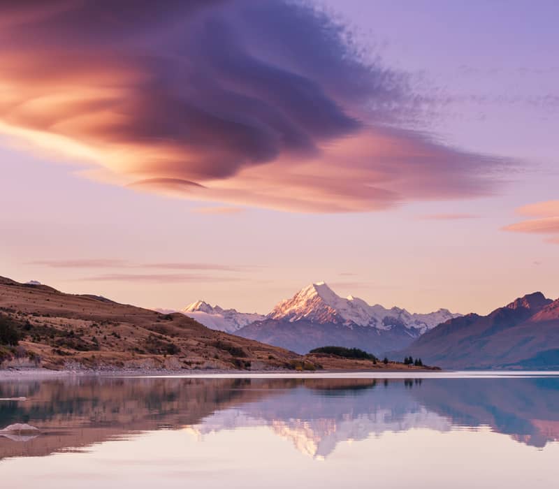 A scenic wide shot of the snow-capped peak of Aoraki Mount Cook reflected perfectly in the calm, mirror-like waters of Lake Pukaki, under a striking pink and purple twilight sky filled with dramatic lenticular cloud formations.