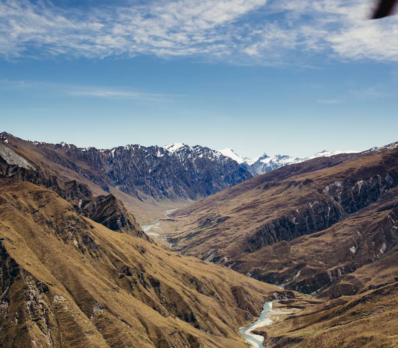 An aerial view over vast, golden-brown mountains with a winding river in a deep valley, leading toward distant snow-capped peaks under a blue sky.