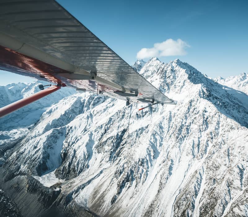 View of snowy mountain peaks from airplane with wing visible under blue sky.