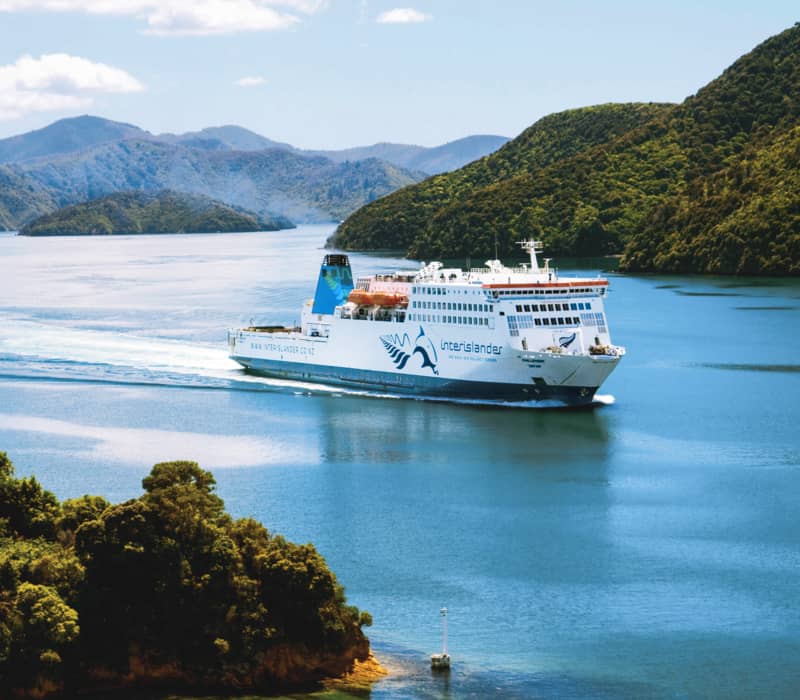 Ferry cruising through blue waters with green forested islands and mountains in the background.