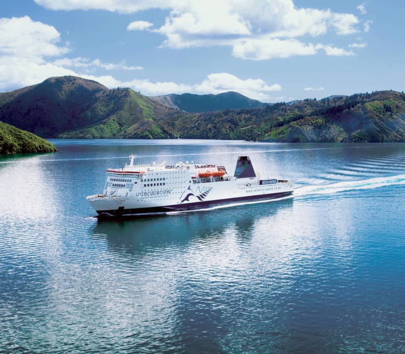 Ferry sailing on calm water with mountains and blue sky in the background.