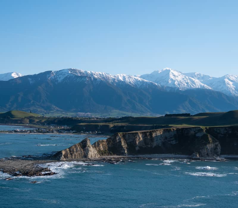 A high-angle coastal view featuring sharp white limestone cliffs jutting into a turquoise ocean, with rolling green hills and a vast range of snow-capped mountains in the background under a clear blue sky.