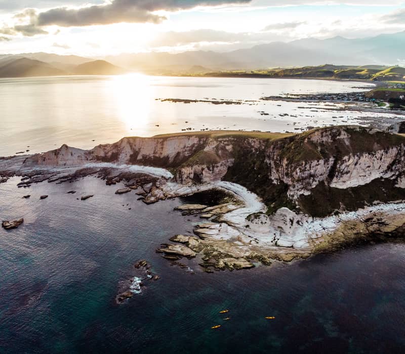 A high-altitude drone view of white limestone cliffs and a rocky reef jutting into a dark teal ocean, with three tiny yellow kayaks in the water and a bright sun glare on the horizon.