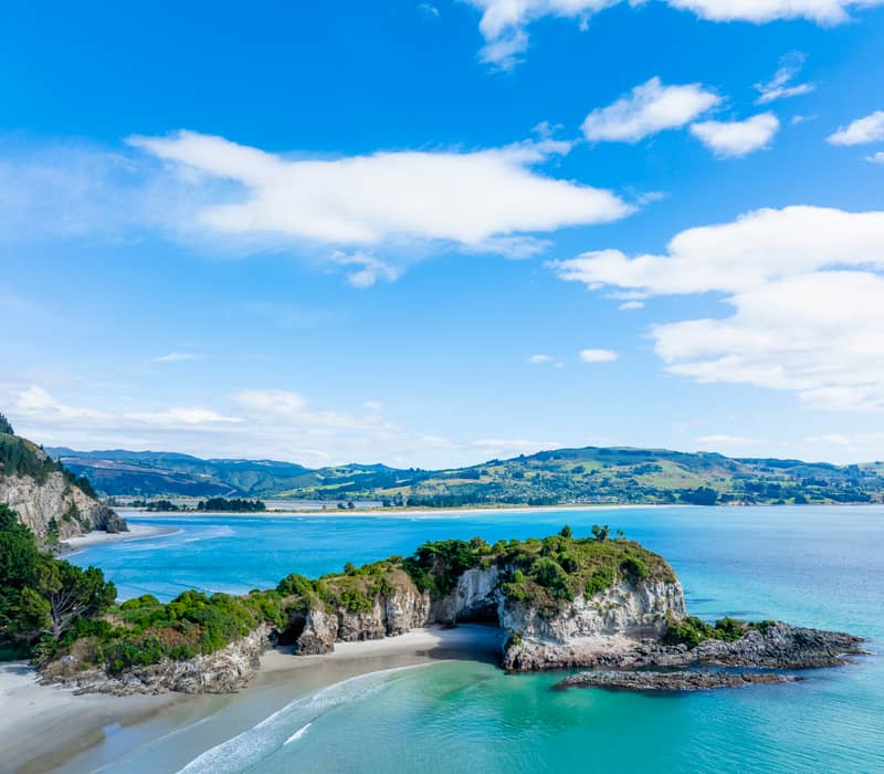 A close aerial drone view of a green, rocky headland with a large sea cave, connected by a narrow white sand beach to a turquoise bay under a bright blue sky.