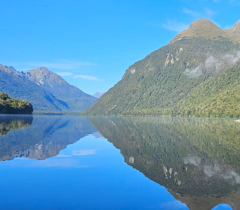 Calm lake reflecting green mountains and blue sky with two people standing by the shore