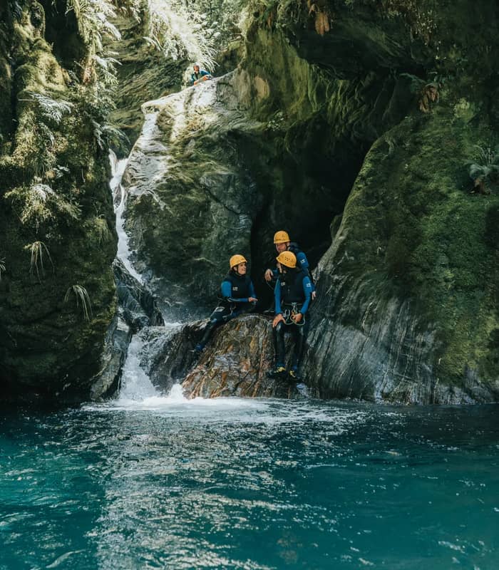 Three people in wetsuits and helmets canyoning at a waterfall in a green rocky gorge with blue water.