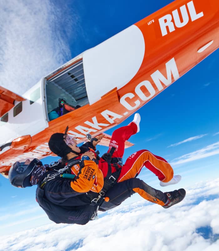Two people in skydiving gear jumping out of an orange and white airplane above clouds.