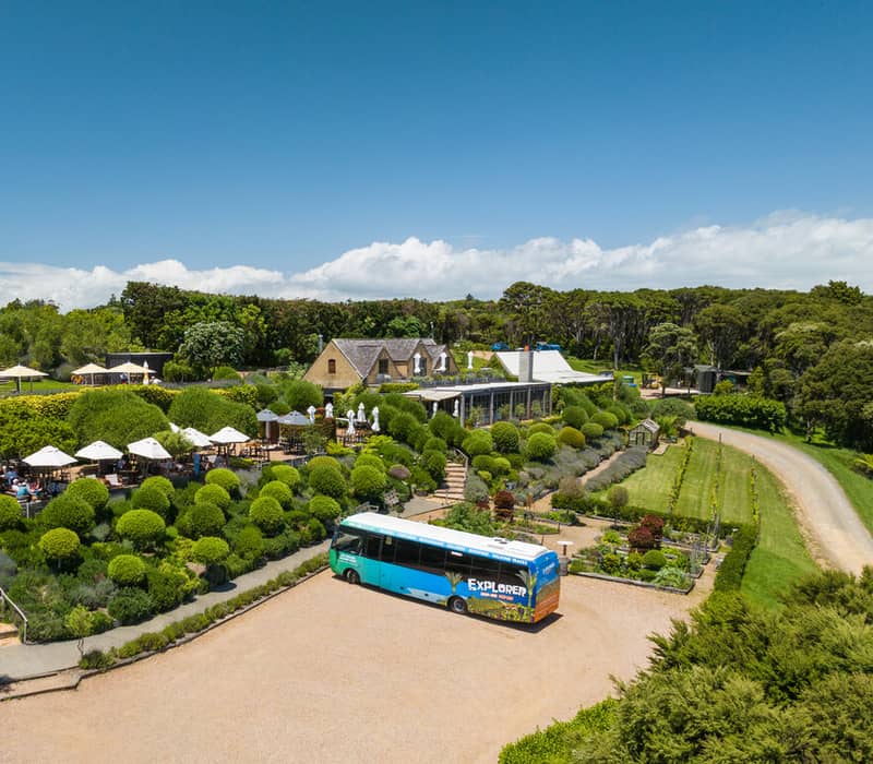 Tour bus parked near a garden with outdoor seating and green bushes under sunny, clear blue skies.
