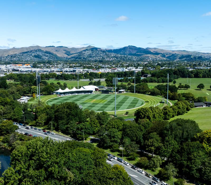 A high-altitude aerial view of a circular green cricket ground with a modern pavilion, surrounded by dense green forest and parkland, with a city and mountains in the background.