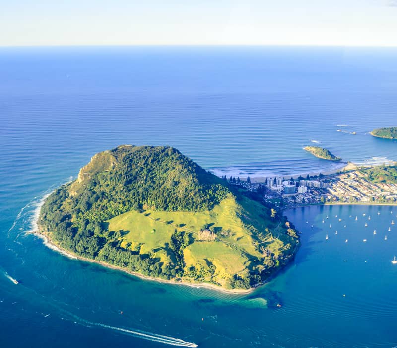 A high-altitude aerial shot of Mauao (Mount Maunganui) bathed in afternoon sunlight, showing the green volcanic cone, the white sand isthmus, and the turquoise waters of the Bay of Plenty filled with boats.