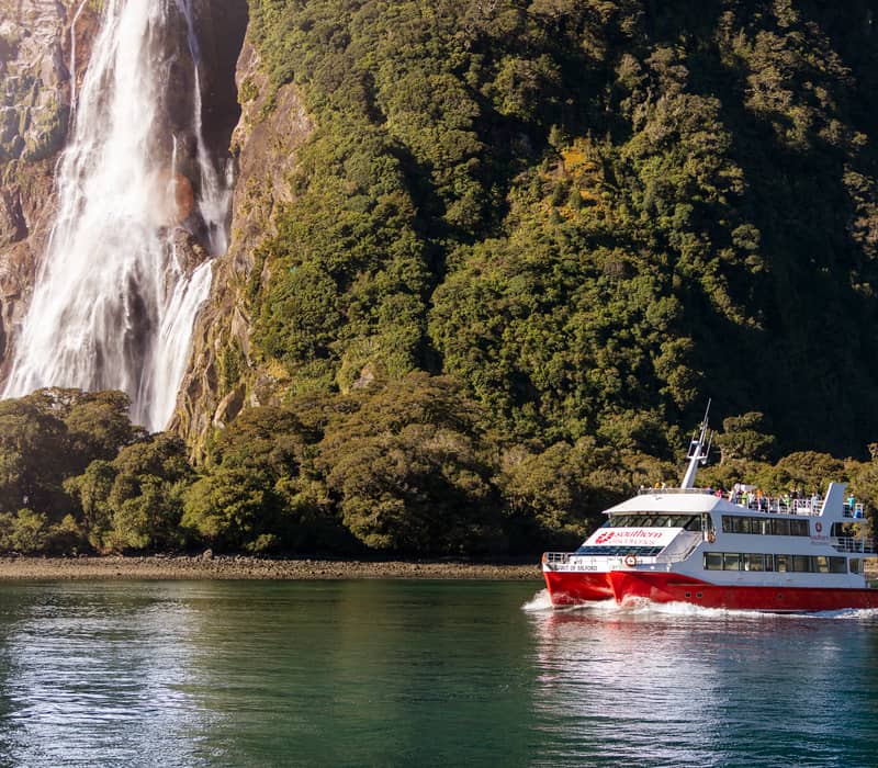 Red and white cruise boat near a waterfall and forested mountain in Milford Sound