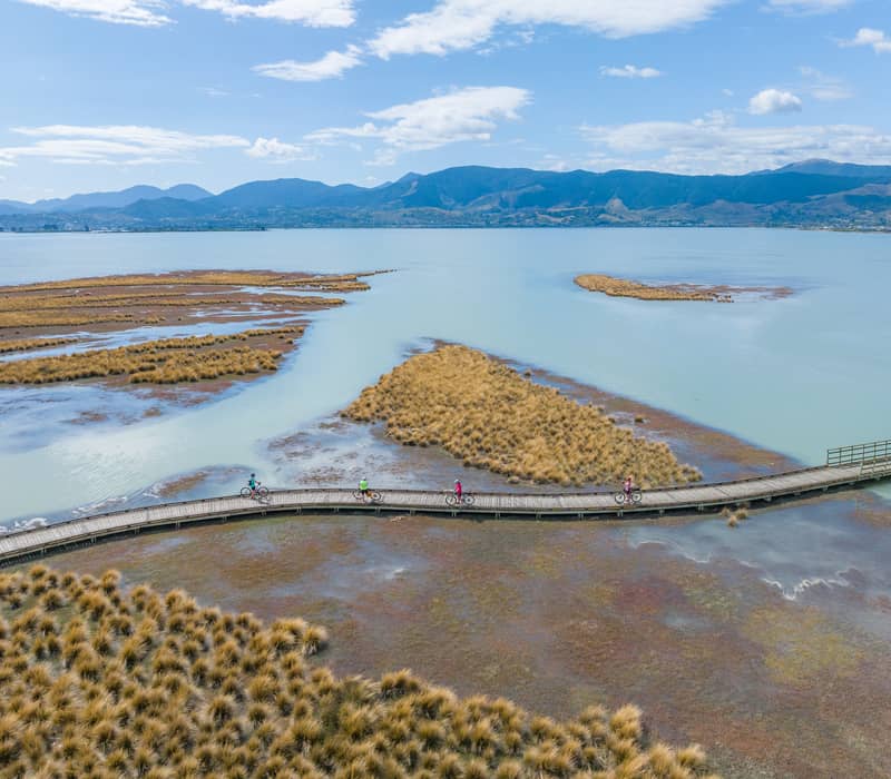 An aerial view of four cyclists riding across a long wooden boardwalk over a shallow coastal inlet with golden marsh grasses and distant blue mountains.