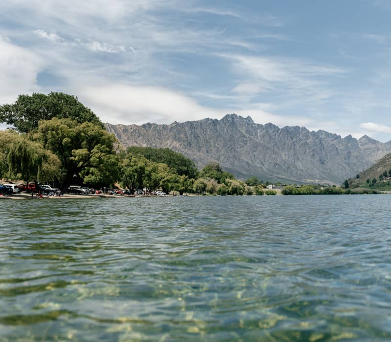 A wide, low-angle shot from the clear, rippling waters of Frankton Beach, with leafy willow trees lining the shore, parked cars, and the jagged, snow-capped Remarkables mountain range in the background.