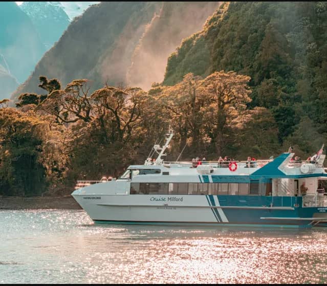 Cruise boat on water near forested mountains with sunlight reflecting on the water.