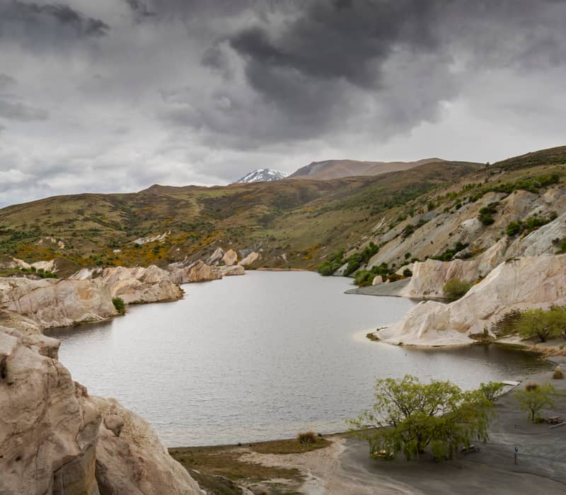 A wide view of Blue Lake in St Bathans, surrounded by dramatic white quartz cliffs and golden-brown hills under a dark, stormy sky in Central Otago.