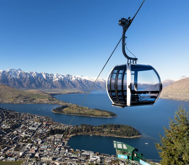 A modern glass gondola cabin suspended high above Queenstown, offering a stunning panoramic view of the blue Lake Wakatipu and the snow-capped Remarkables mountain range.