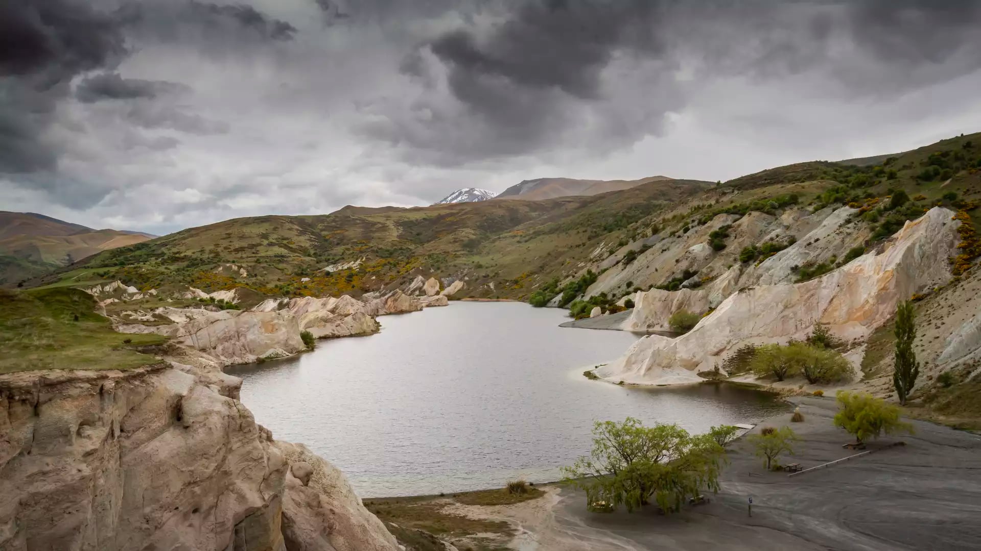 A wide view of Blue Lake in St Bathans, surrounded by dramatic white quartz cliffs and golden-brown hills under a dark, stormy sky in Central Otago.