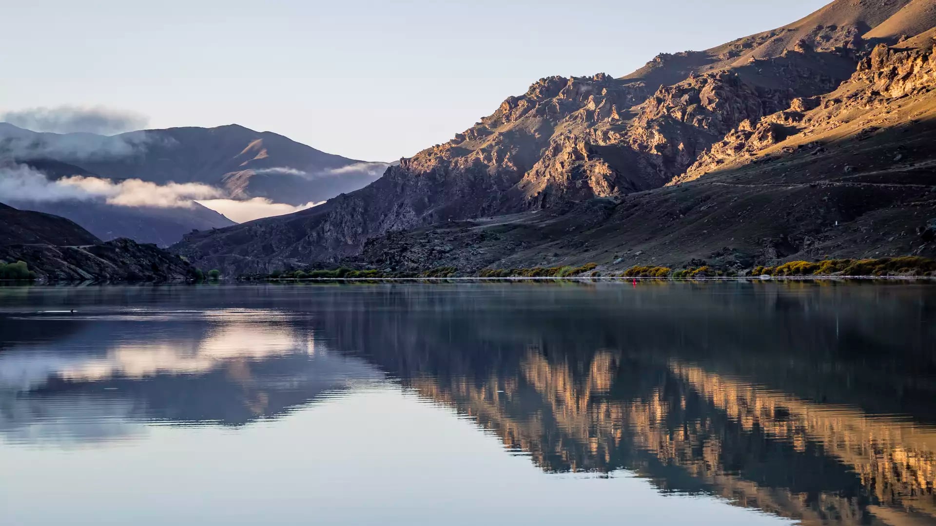 A symmetrical reflection of jagged, sunlit mountains and soft morning mist on the glassy surface of Lake Dunstan in Central Otago.