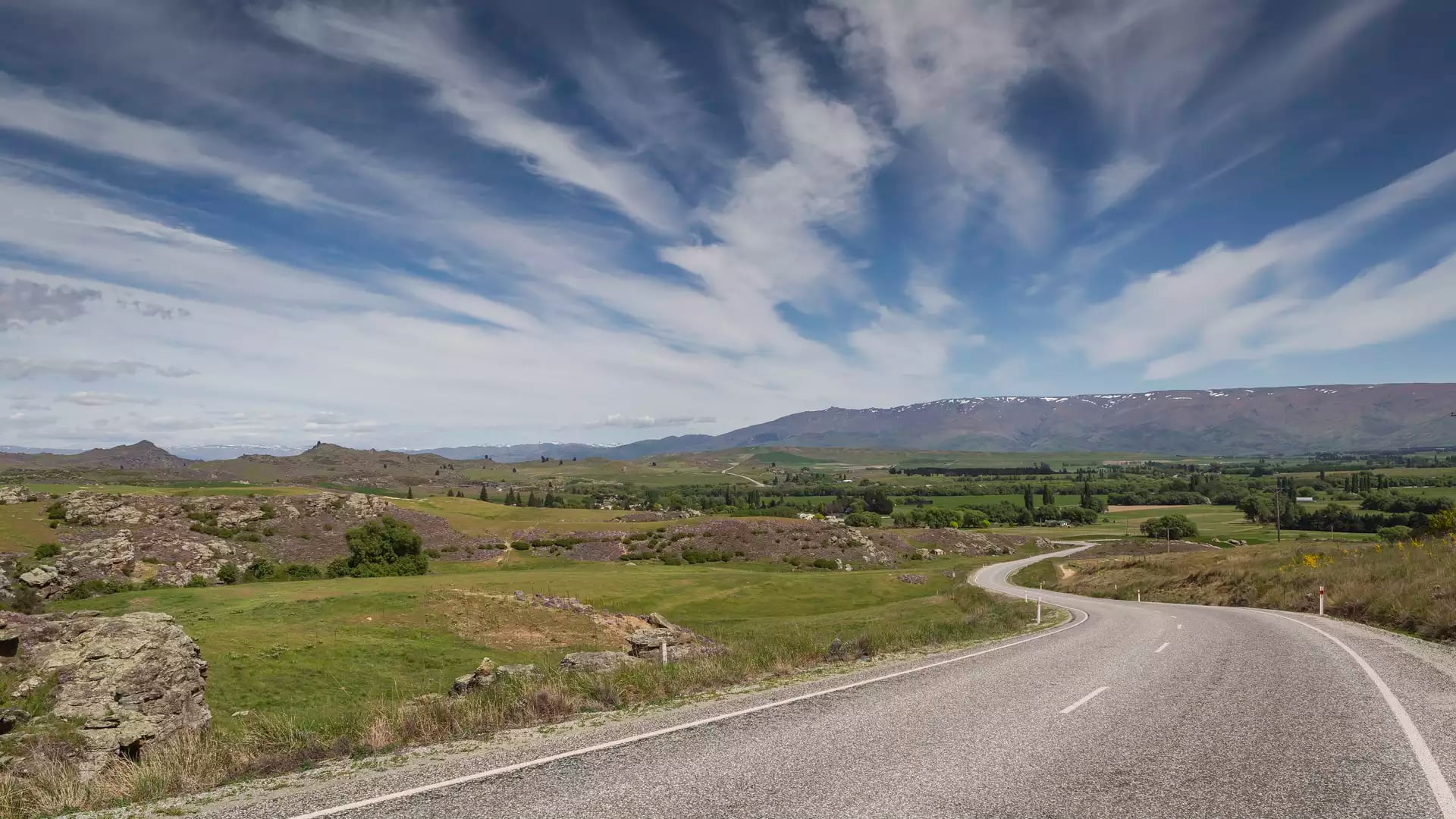 A winding two-lane highway curves through a vast landscape of green fields, rocky schist outcrops, and distant purple mountains under a dramatic blue sky with wispy white clouds.