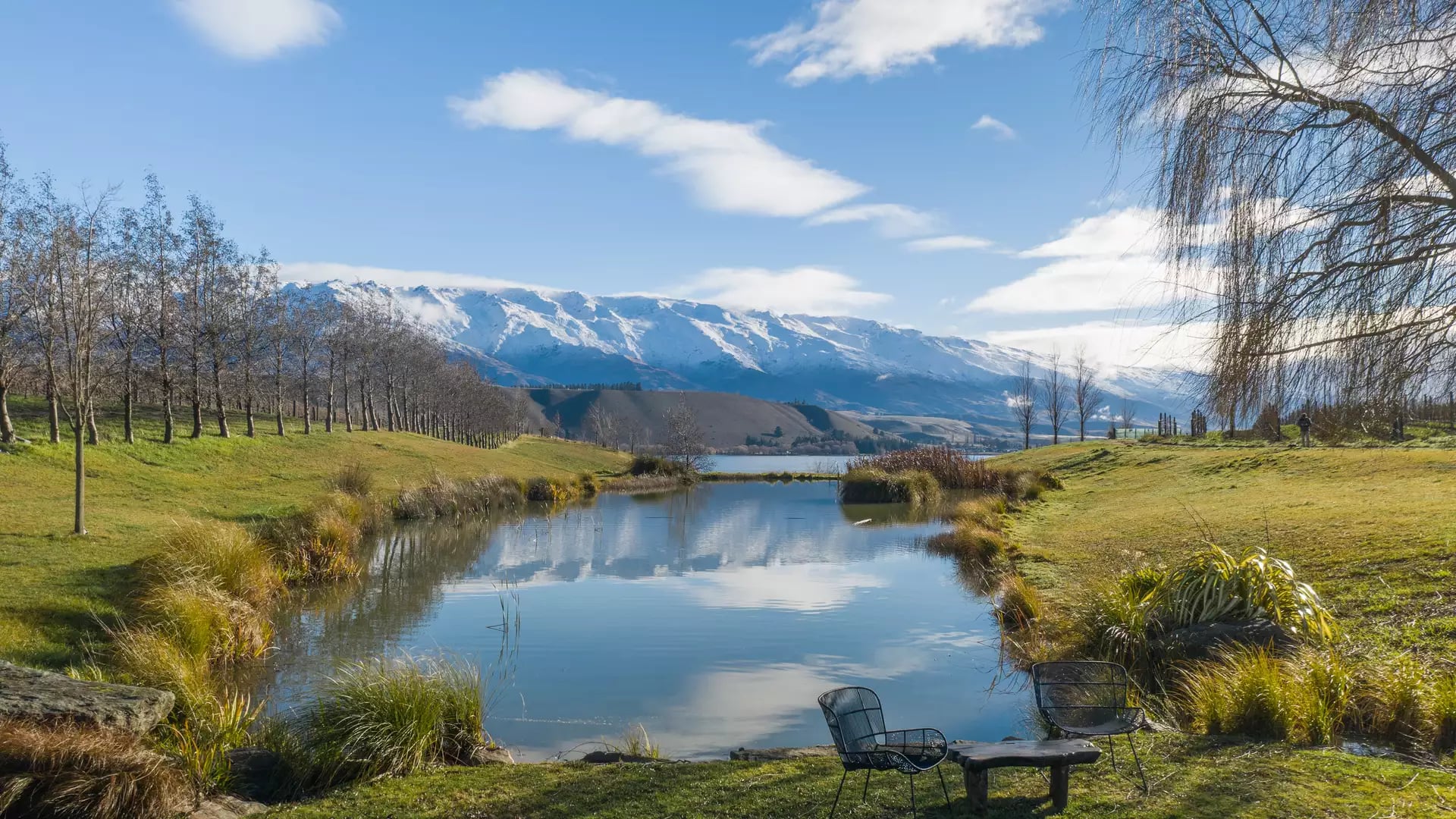 Two black mesh outdoor chairs and a small wooden table sit on a grassy bank overlooking a calm pond that reflects snow-capped mountains and a blue sky at Cloudy Bay vineyard.