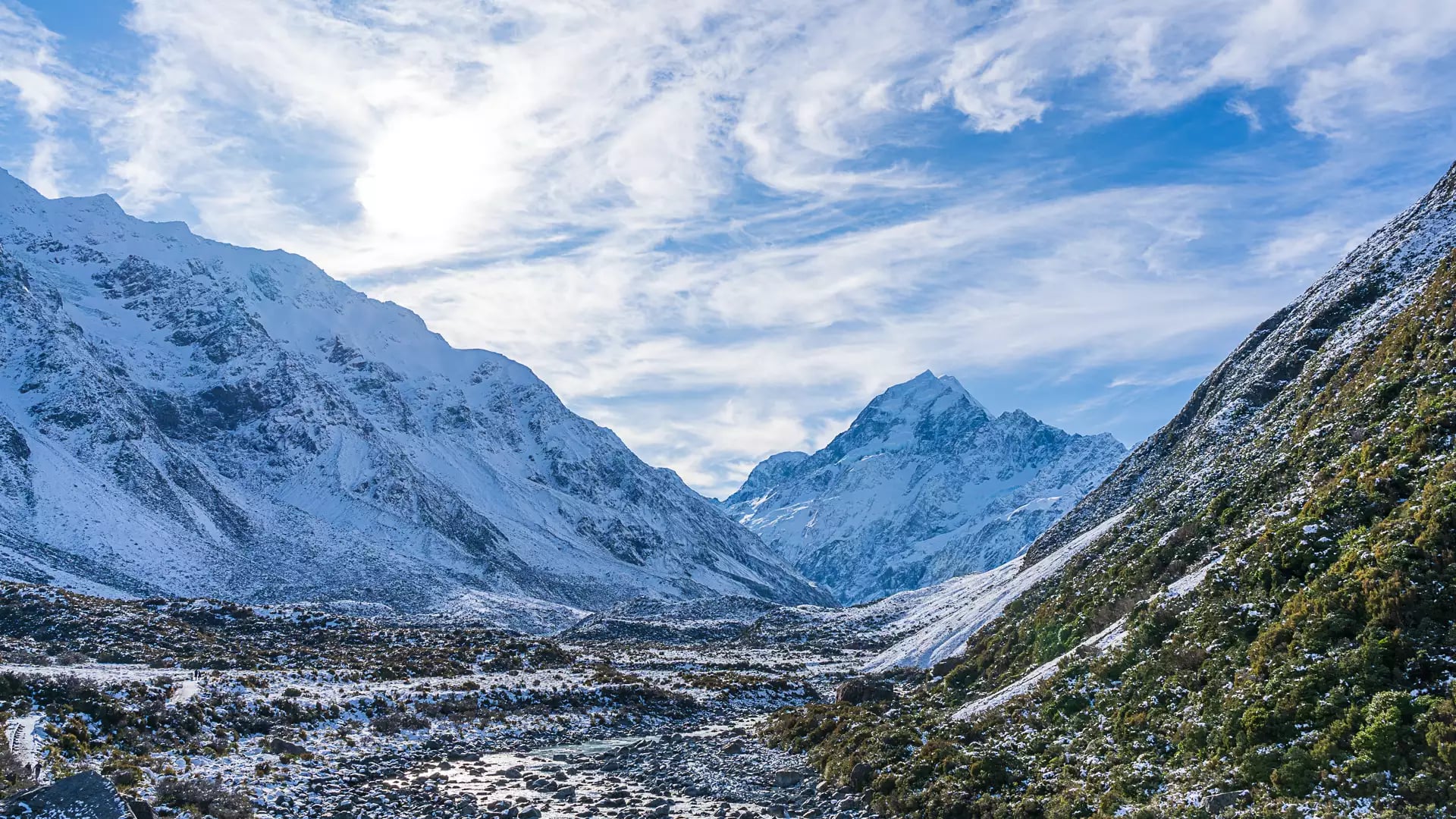 A wide view of the rocky Hooker River valley floor leading toward the massive, snow-covered peak of Aoraki / Mount Cook under a bright, wispy blue sky.