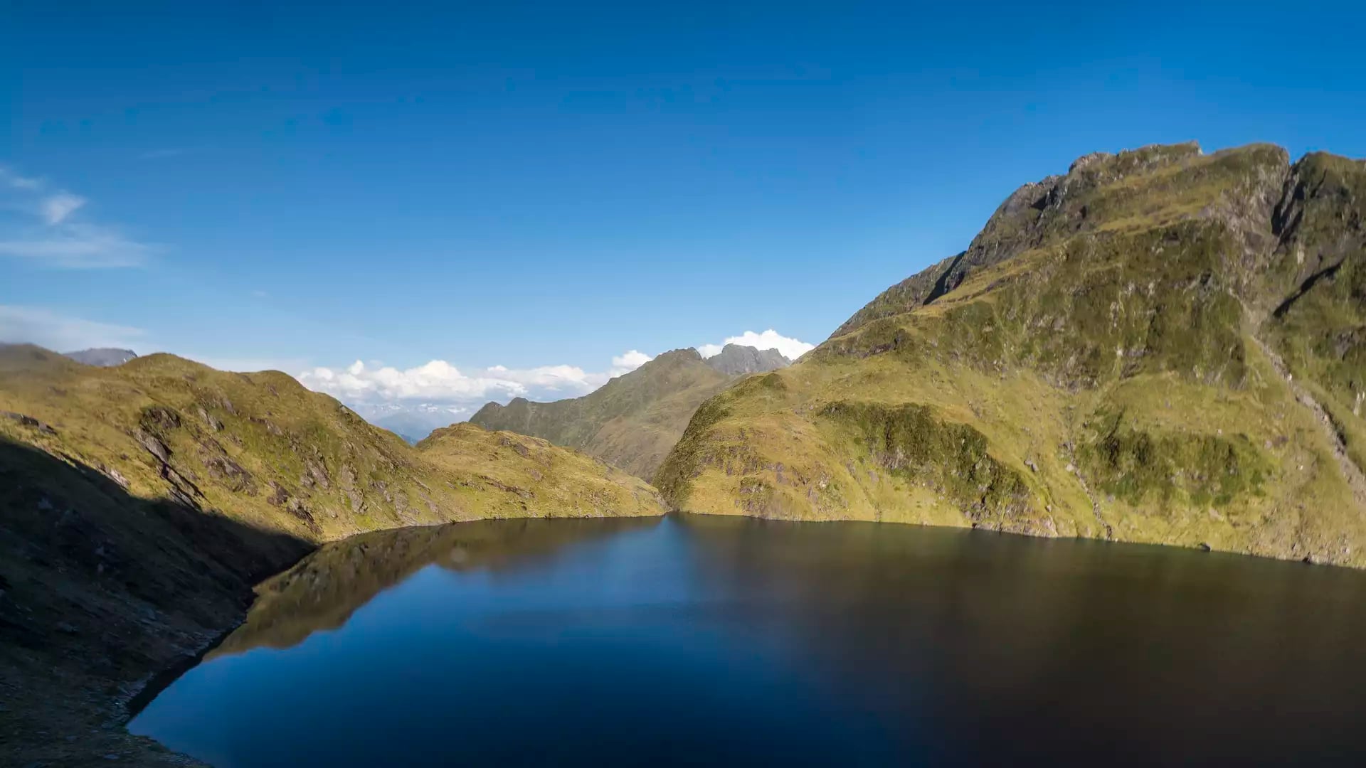 A wide panoramic view of a deep blue alpine lake surrounded by steep, sunlit golden-green mountain ridges under a clear blue sky.