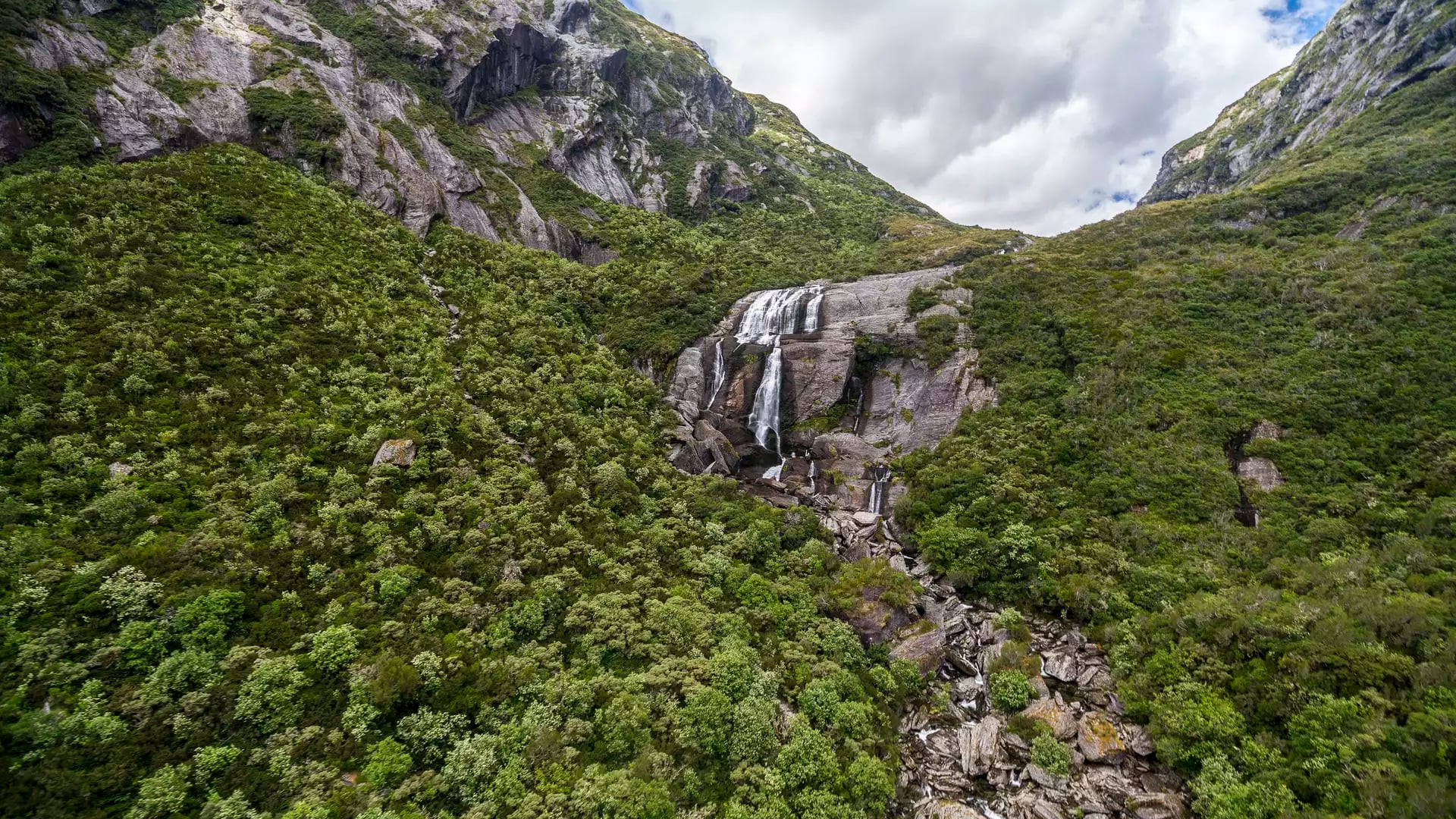A wide waterfall cascades down a steep, rocky mountain face surrounded by dense green native bush in the Haast Pass.