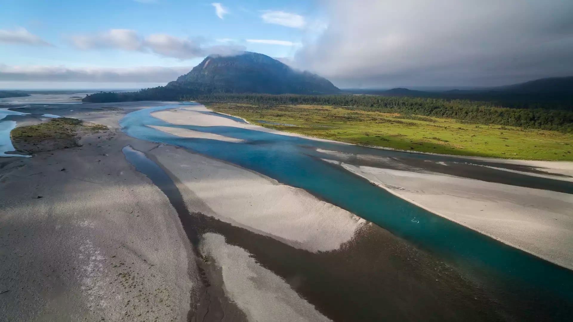 An aerial view of the Haast River’s turquoise braided channels flowing through wide shingle banks toward a forested mountain under a soft, morning sky.