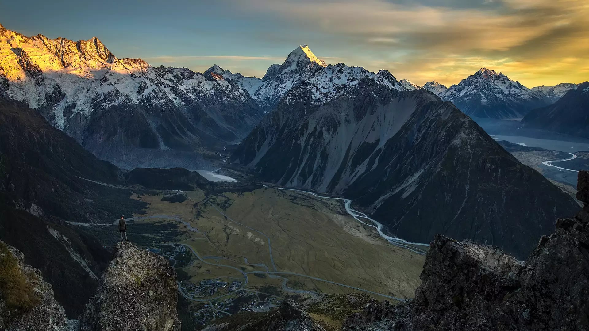 A high-altitude view of a person standing on a rocky outcrop overlooking a vast glacial valley, with the sun-drenched peak of Aoraki / Mount Cook in the distance under a golden sky.