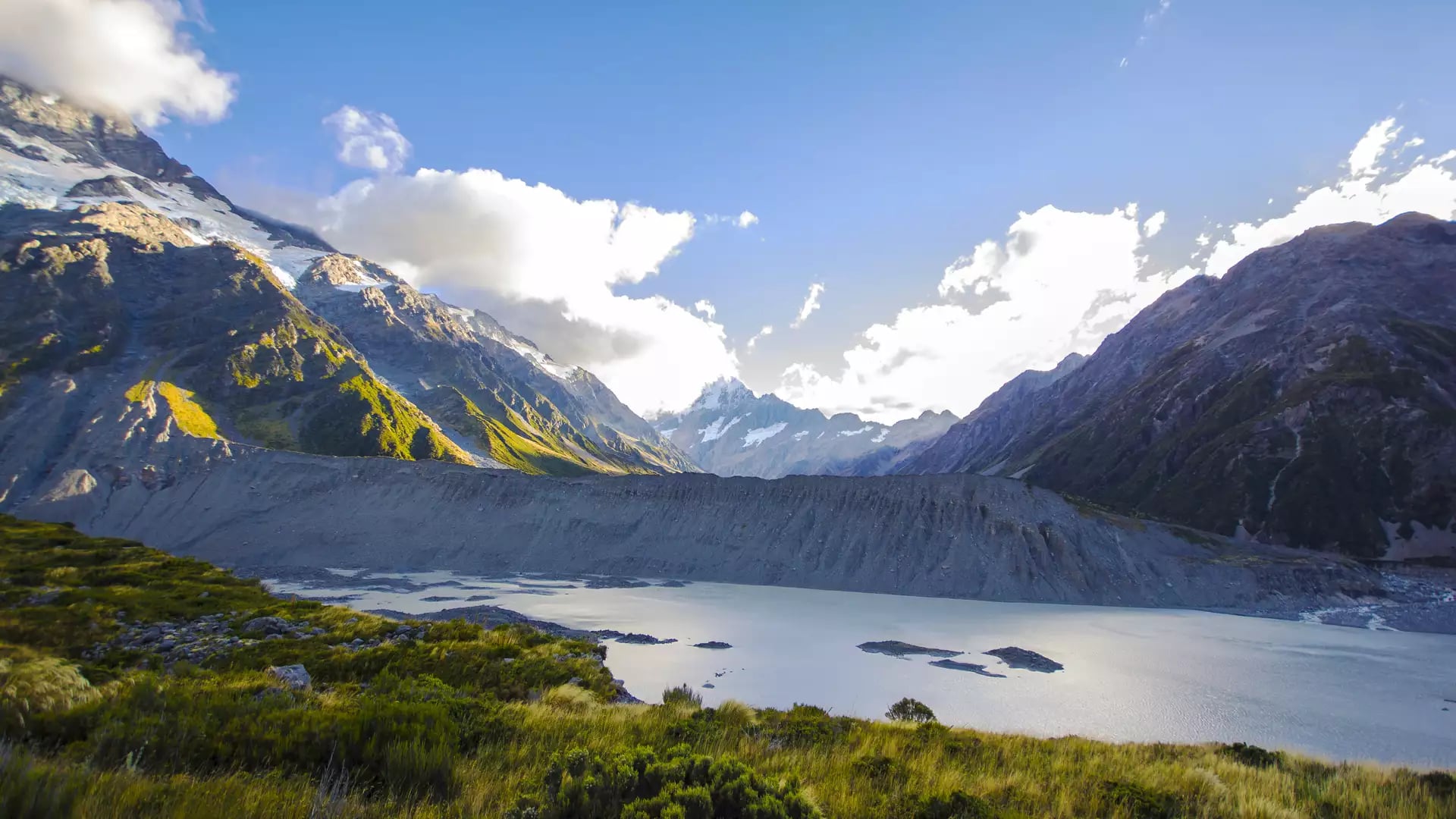 A wide-angle daytime view of the Mueller Glacier lake and moraine wall, with sunlit green and gold tussock in the foreground and snow-capped peaks under a bright, cloudy sky.