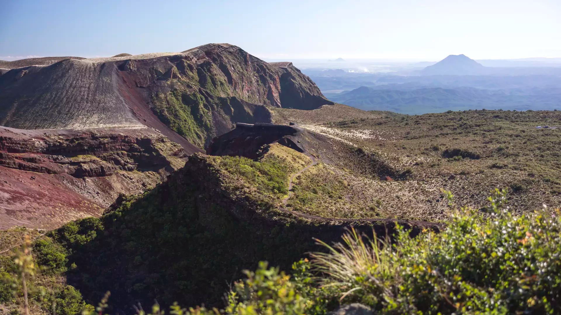 An expansive view of the deep, reddish-brown volcanic crater of Mount Tarawera in Rotorua, with a hiking trail winding along the rim and distant mountains under a clear sky.