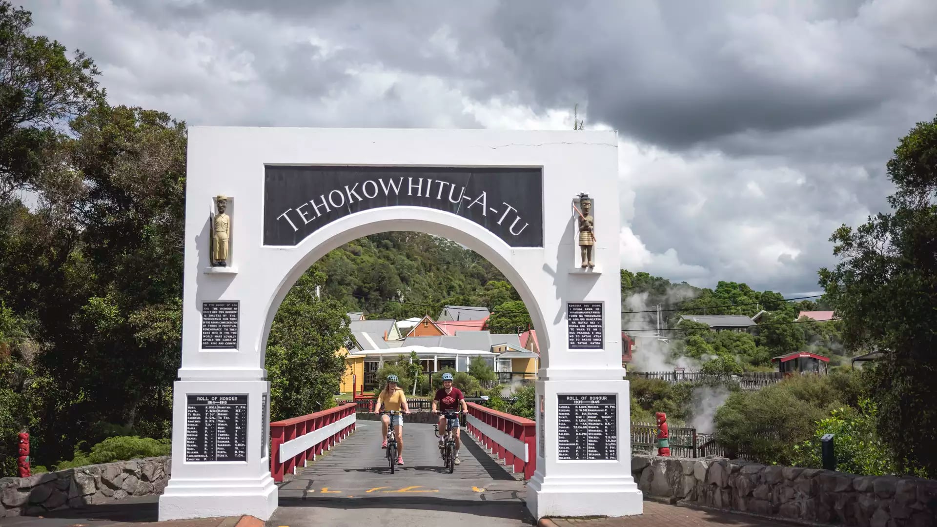 Two cyclists ride through the historic white "Te Hokowhitu-a-Tu" memorial archway into Whakarewarewa Living Māori Village, with geothermal steam visible in the background.