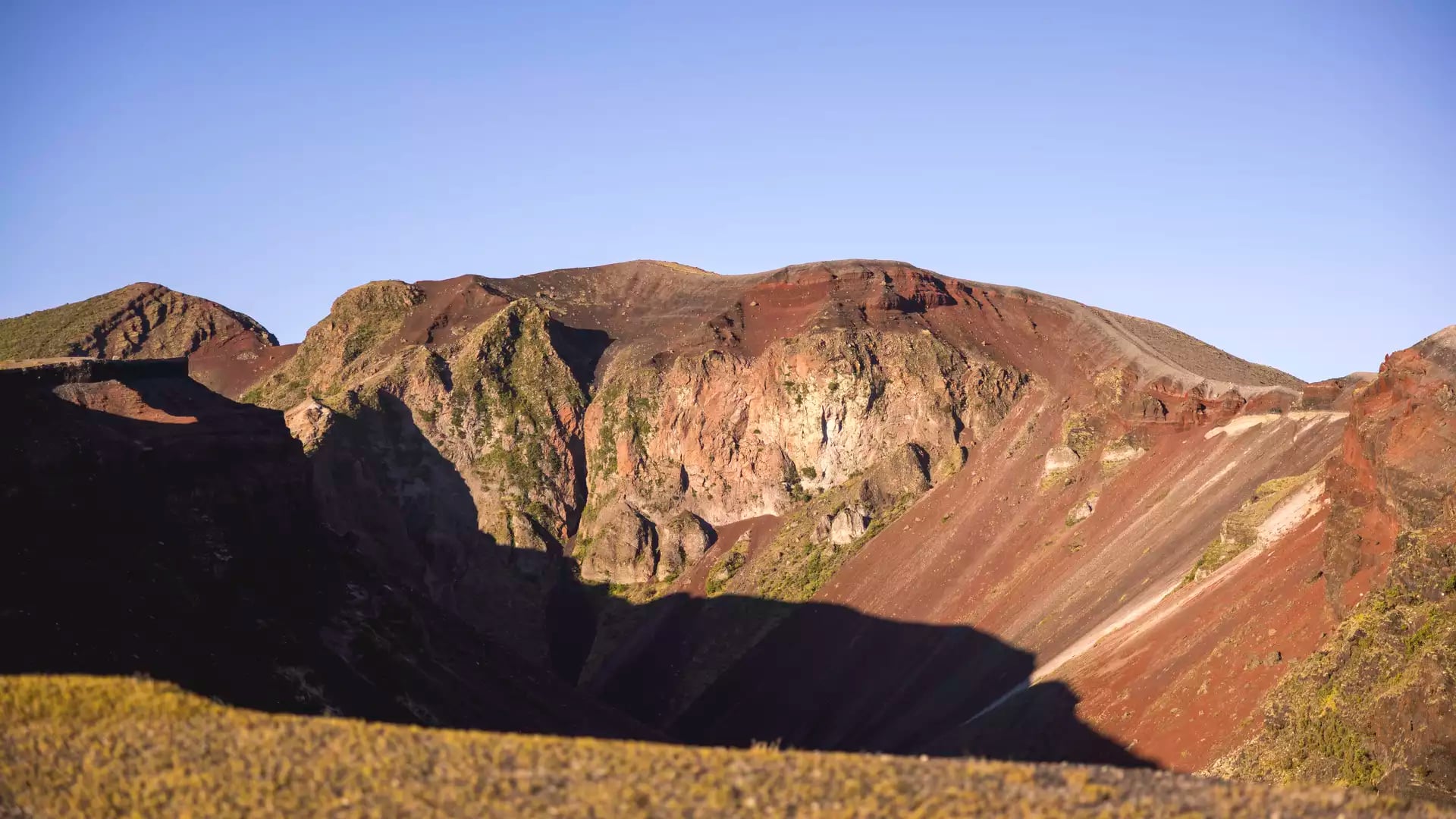 A wide view of the colorful volcanic slopes of Mount Tarawera under a clear sky, showing deep red scoria and rocky ridges in the afternoon sun.