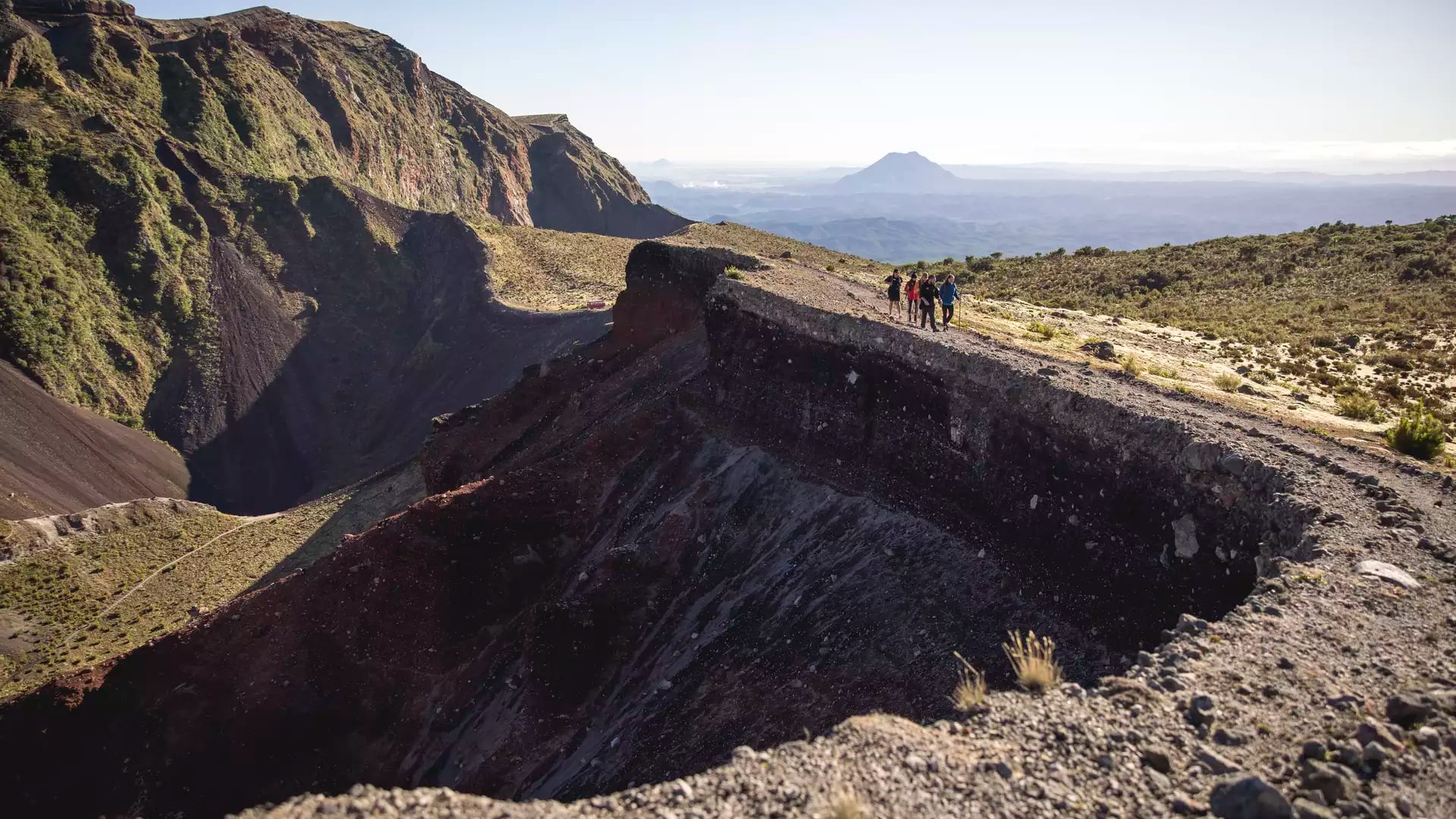 A group of four hikers walks along the narrow, rocky rim of a massive volcanic crater on Mount Tarawera, with deep red and purple chasm walls falling away to the left.