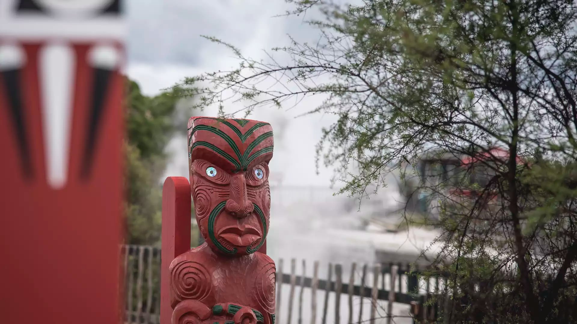 A close-up of a traditional red Māori wood carving with iridescent pawa shell eyes, standing in front of a steaming geothermal field and native manuka trees.