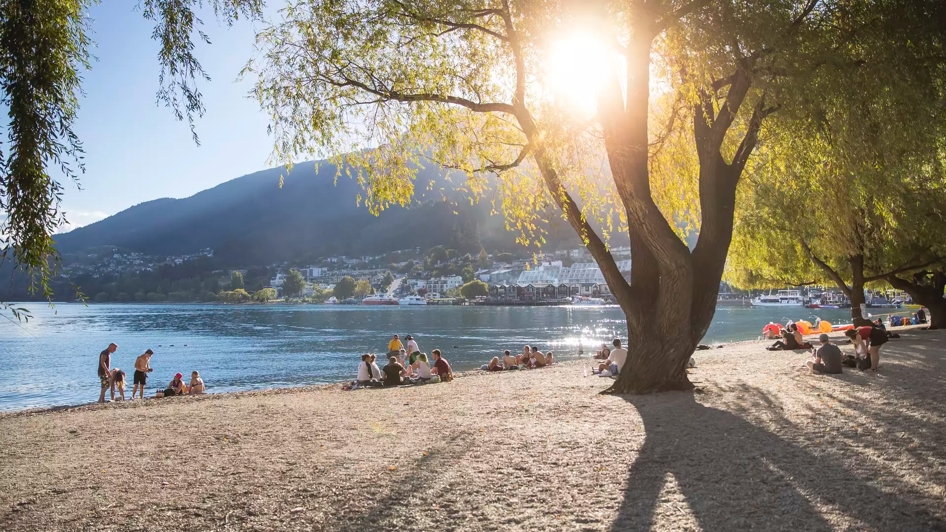 People lounging on the pebbled shore of Queenstown Beach under the shade of large willow trees, with the sun shining brightly over Lake Wakatipu and the surrounding mountains.