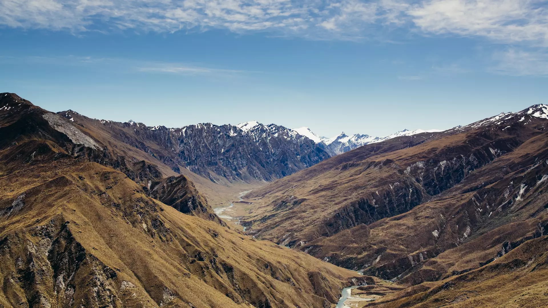 An aerial view over vast, golden-brown mountains with a winding river in a deep valley, leading toward distant snow-capped peaks under a blue sky.