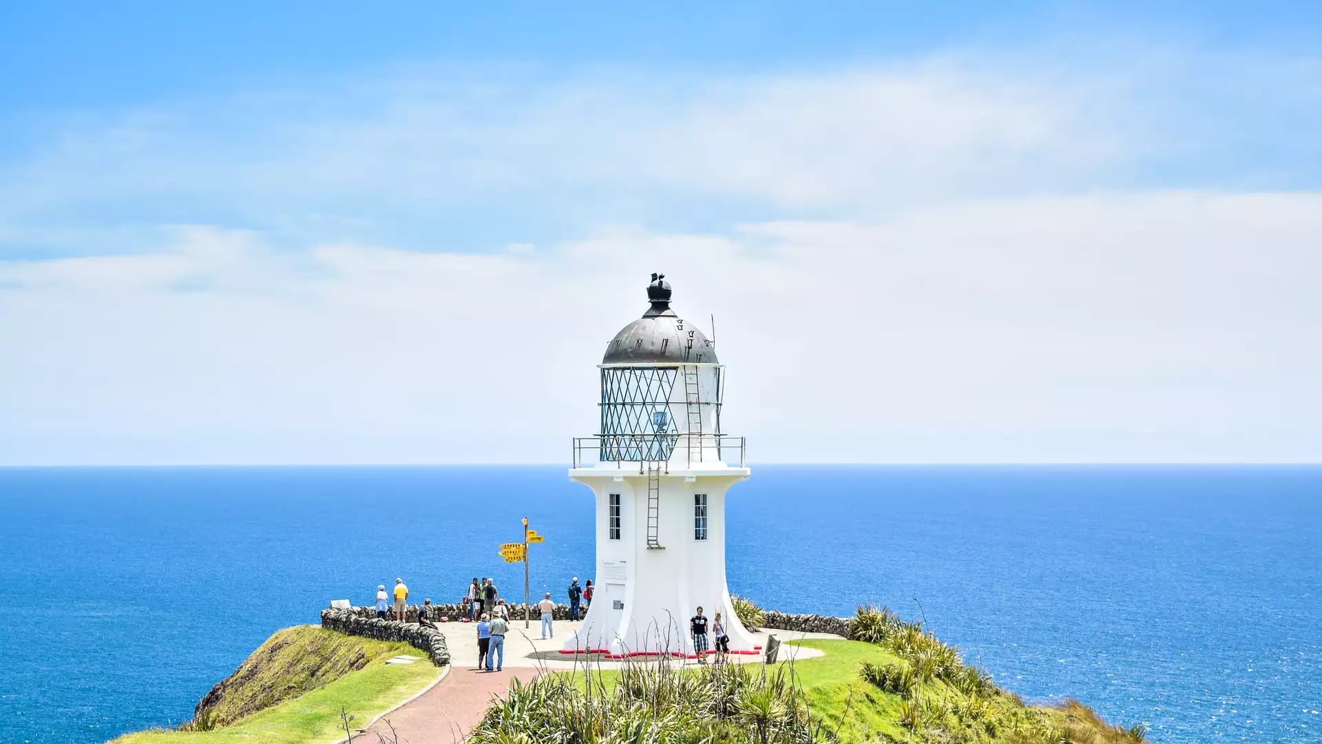 White lighthouse on cliff edge with people and vast blue ocean under a partly cloudy sky