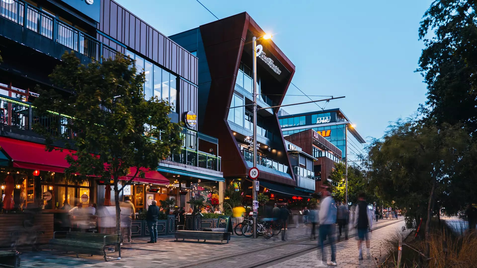 A busy street scene at dusk on Oxford Terrace in Christchurch, featuring glowing restaurants and modern architecture.