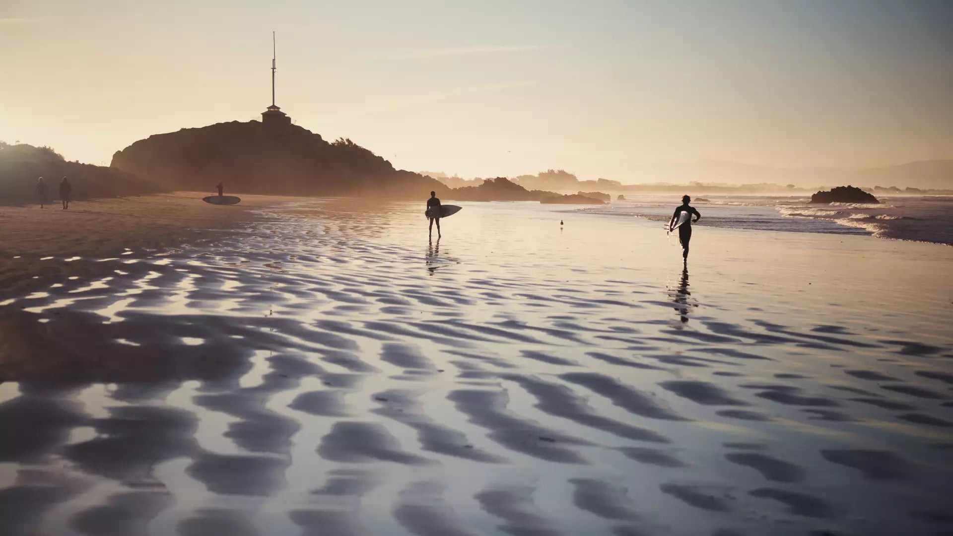 Silhouetted surfers walking on rippled wet sand at Sumner Beach during a golden sunset, with the dark shape of Cave Rock in the distance.