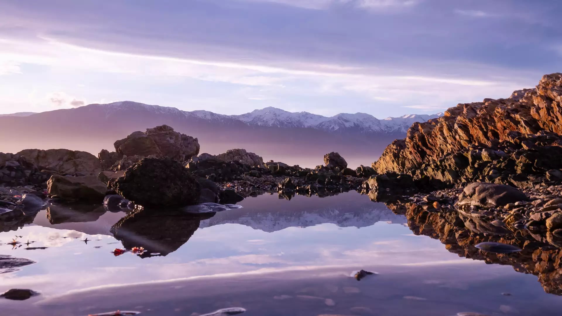A calm coastal rock pool reflects snow-capped mountains under a soft purple and pink sunset sky.