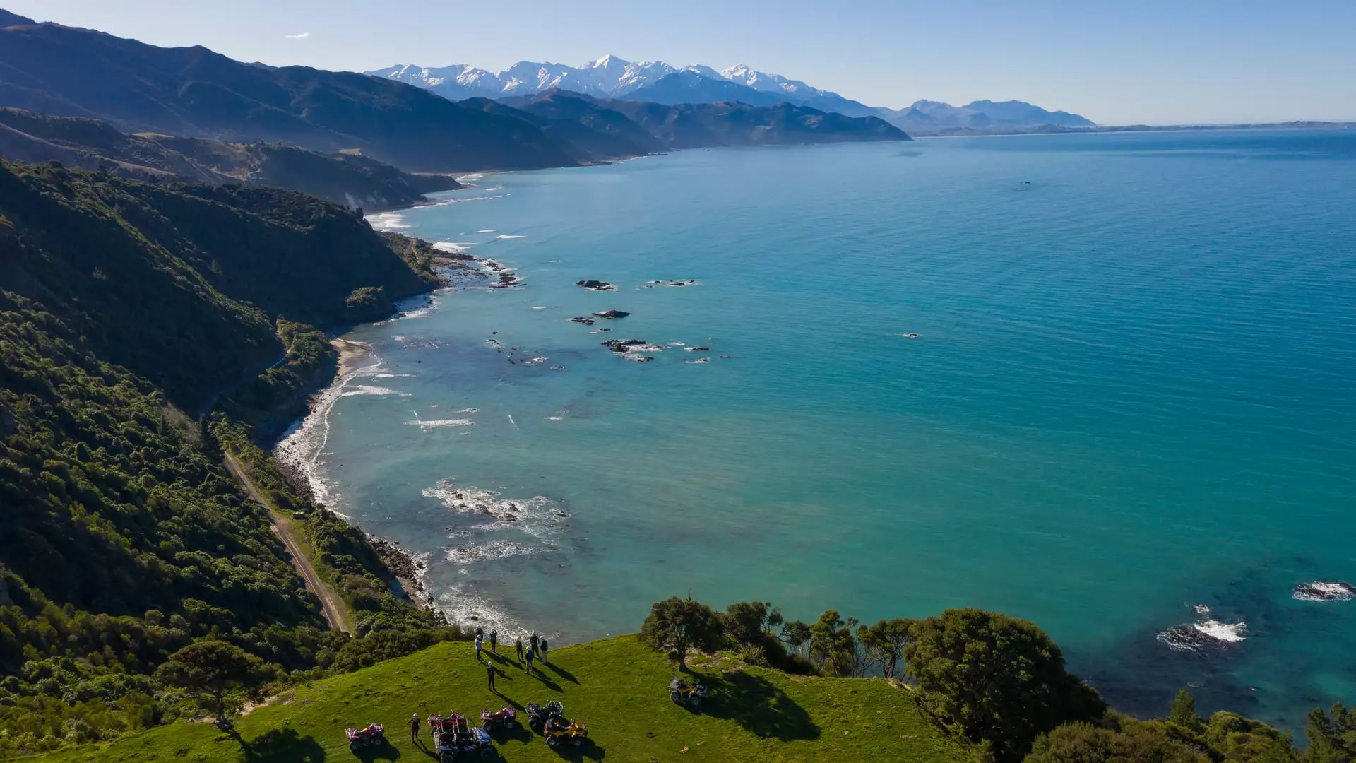 An aerial view of a group of people and quad bikes on a vibrant green grassy cliff overlooking a rugged coastline with turquoise water and distant snow-capped mountains.